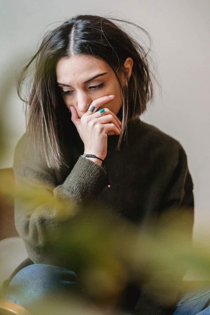 Sad young woman in casual outfit crying with hand on face while sitting on wooden chair in light flat